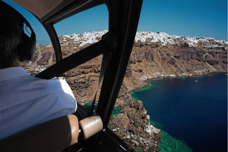 View from inside a helicopter flying over a coastal caldera landscape, with volcanic cliffs, white buildings, and deep blue sea visible below.