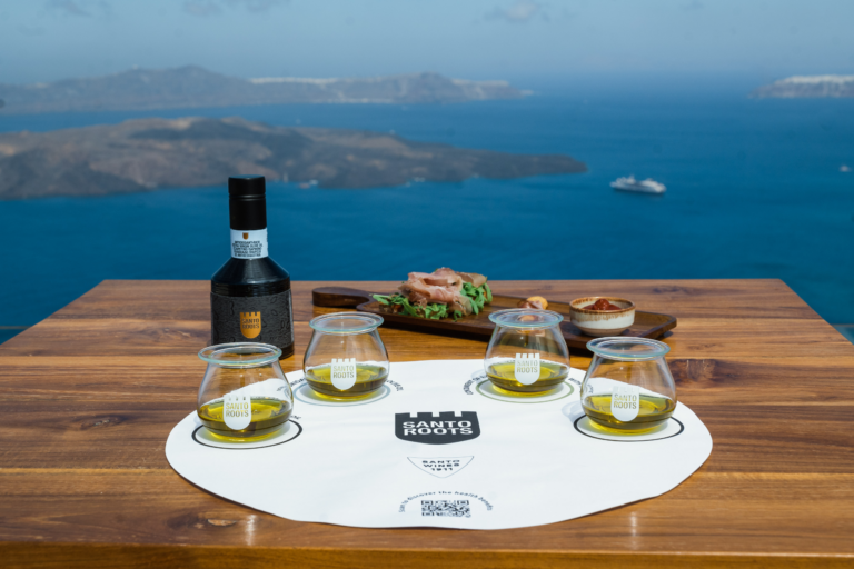 Four glasses of golden extra virgin olive oil on a Santo Roots tasting mat with a bottle of Santo Roots olive oil, charcuterie board, and panoramic views of the Santorini caldera and Aegean Sea.