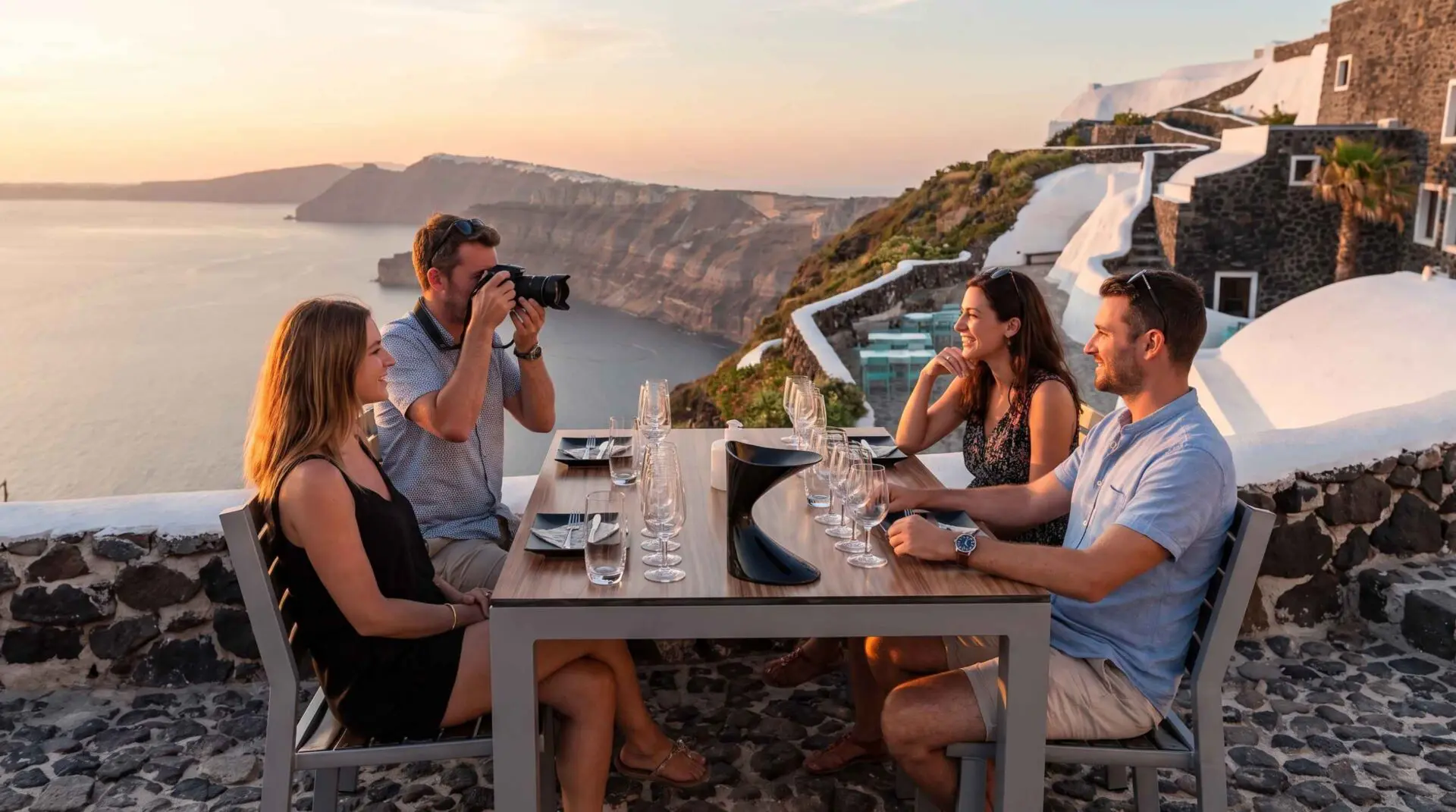 Two couples at a caldera-view restaurant table in Santorini at sunset, wine glasses set for a tasting while one guest photographs the view.