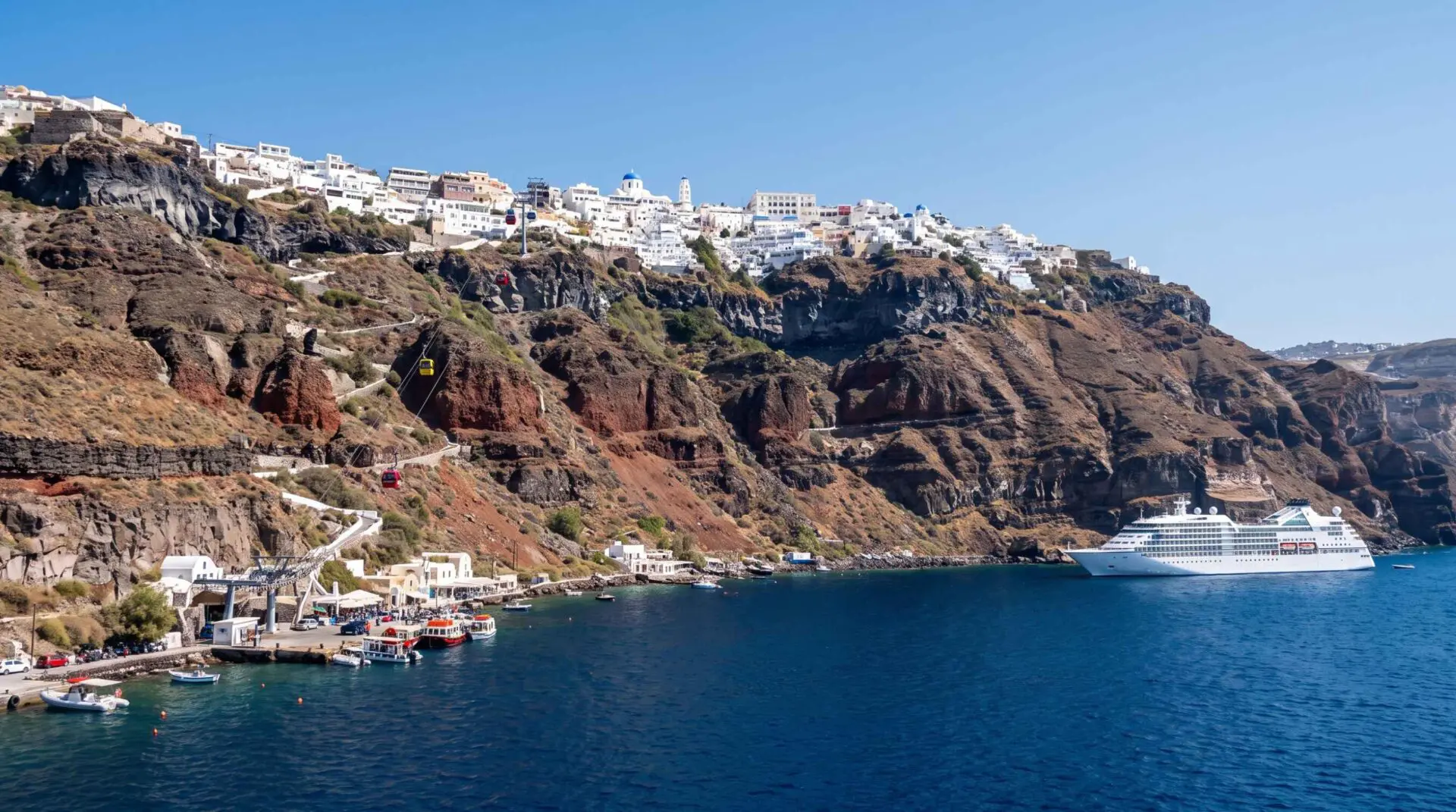 The Santorini cable car climbing the caldera cliff to Fira above the Old Port, with boats below and a cruise ship anchored in the blue caldera.