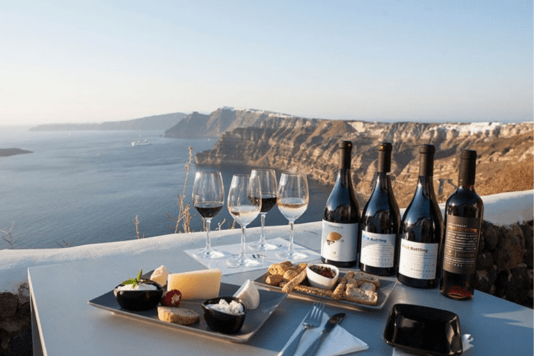 Santorini private wine tour: Bottles of Santorini wine and glasses, accompanied by cheese and bread, overlook a stunning caldera view.