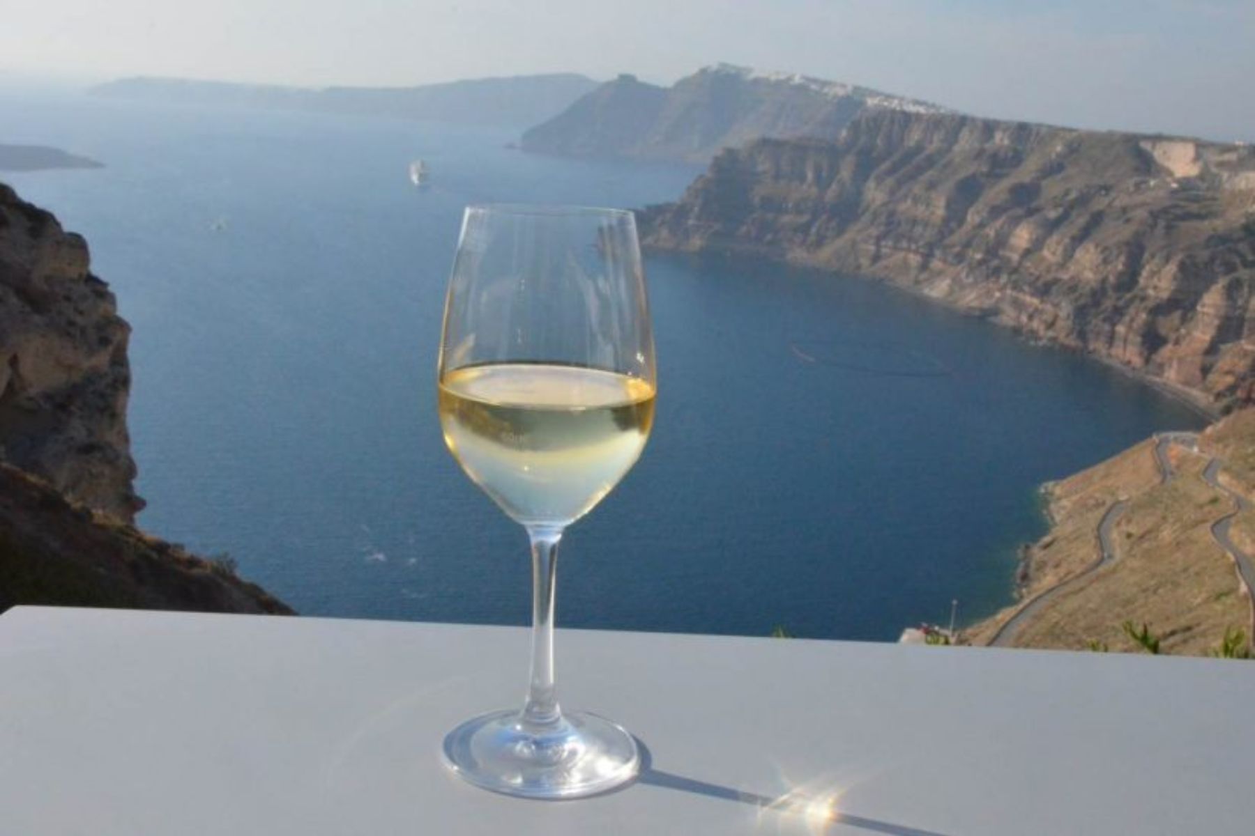 Santorini Wine Tasting with a View A glass of white wine sits on a table overlooking the Aegean Sea and the volcanic cliffs of Santorini, Greece.