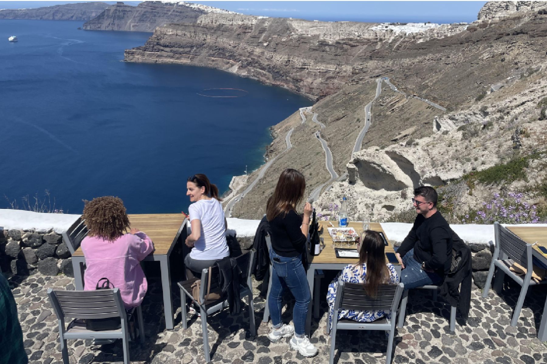 Santorini Wine Tour with a View People are seated at two tables outdoors, enjoying a meal and drinks with a stunning view of the Santorini caldera.