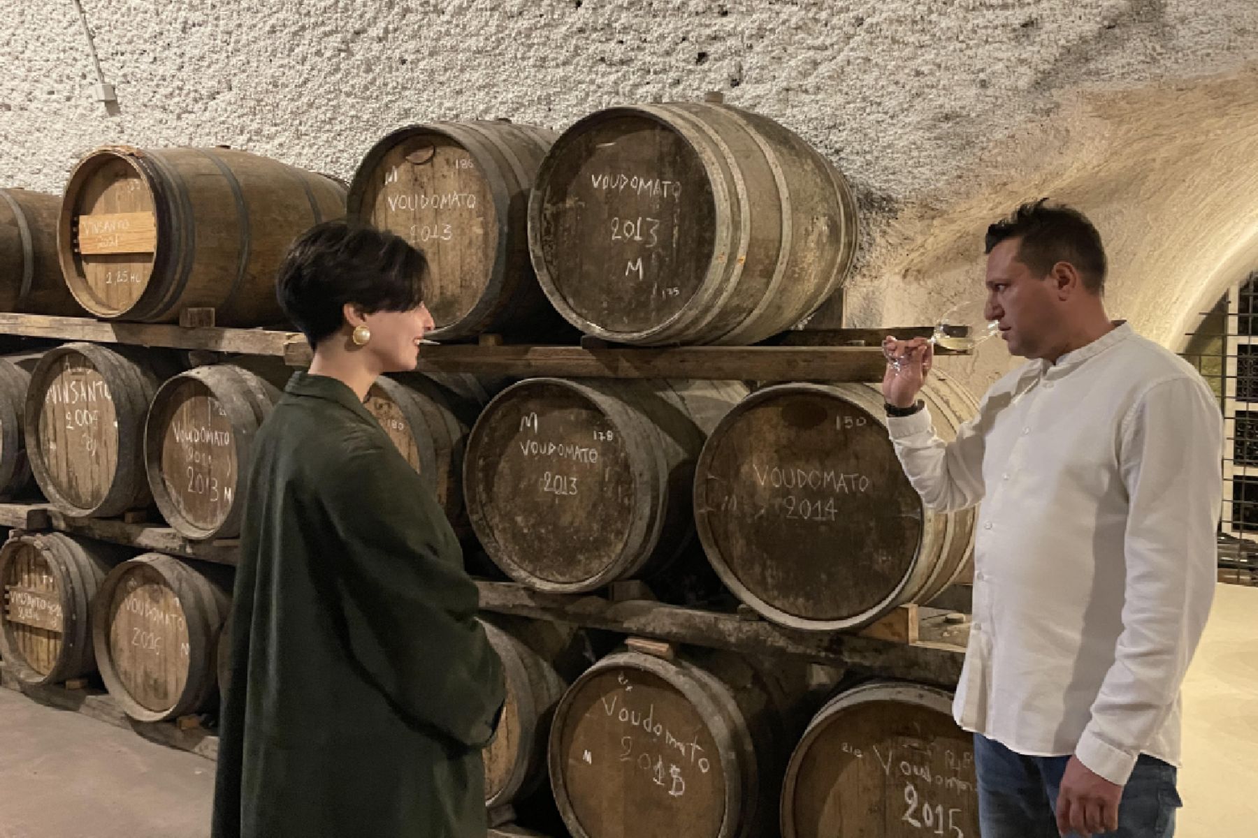 Santorini Wine Tour Two people on a Santorini wine tour sample wine in a cellar surrounded by oak barrels.