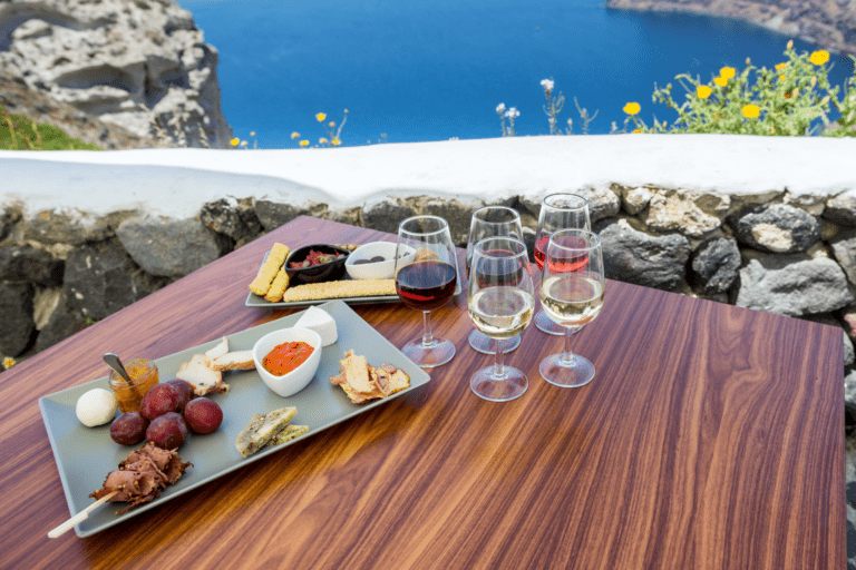 A wooden table set for a private daytime wine tour in Santorini overlooks the Aegean Sea. Wine glasses filled with white and red wine are arranged alongside platters of cheese, meats, and bread.