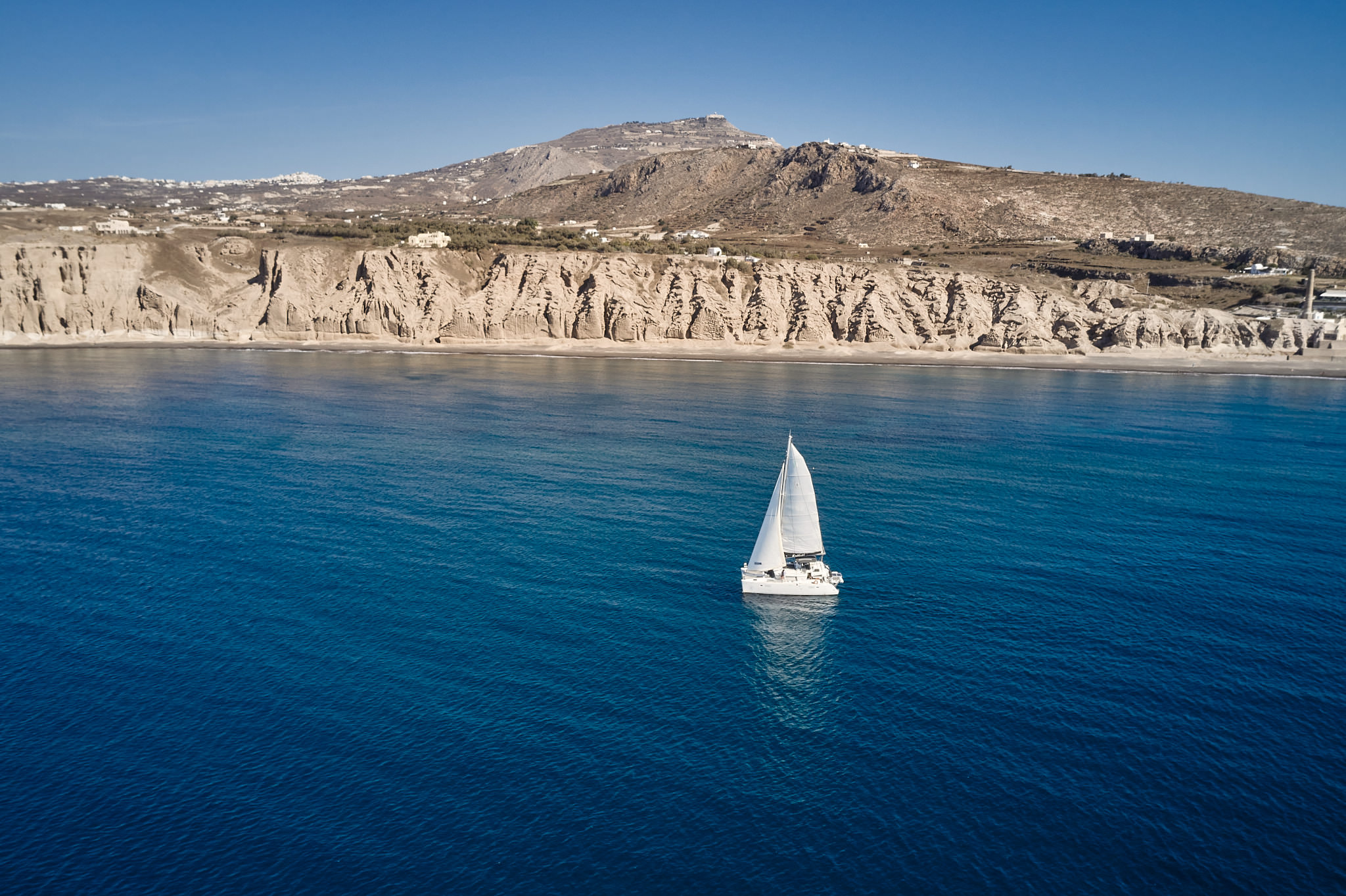 Private Sailing Cruise along the Coast of Santorini Sailing catamaran cruising near the coastline of Santorini with rugged cliffs and clear blue waters in the background.