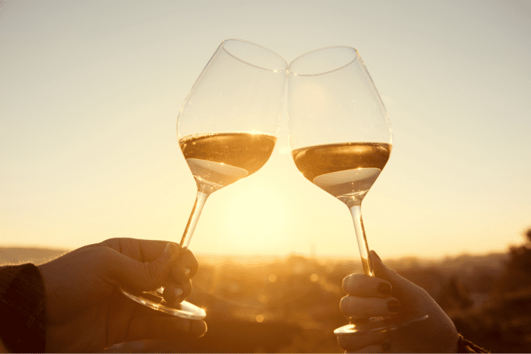 Two people clink glasses of white wine at sunset, silhouetted against a warm golden sky.