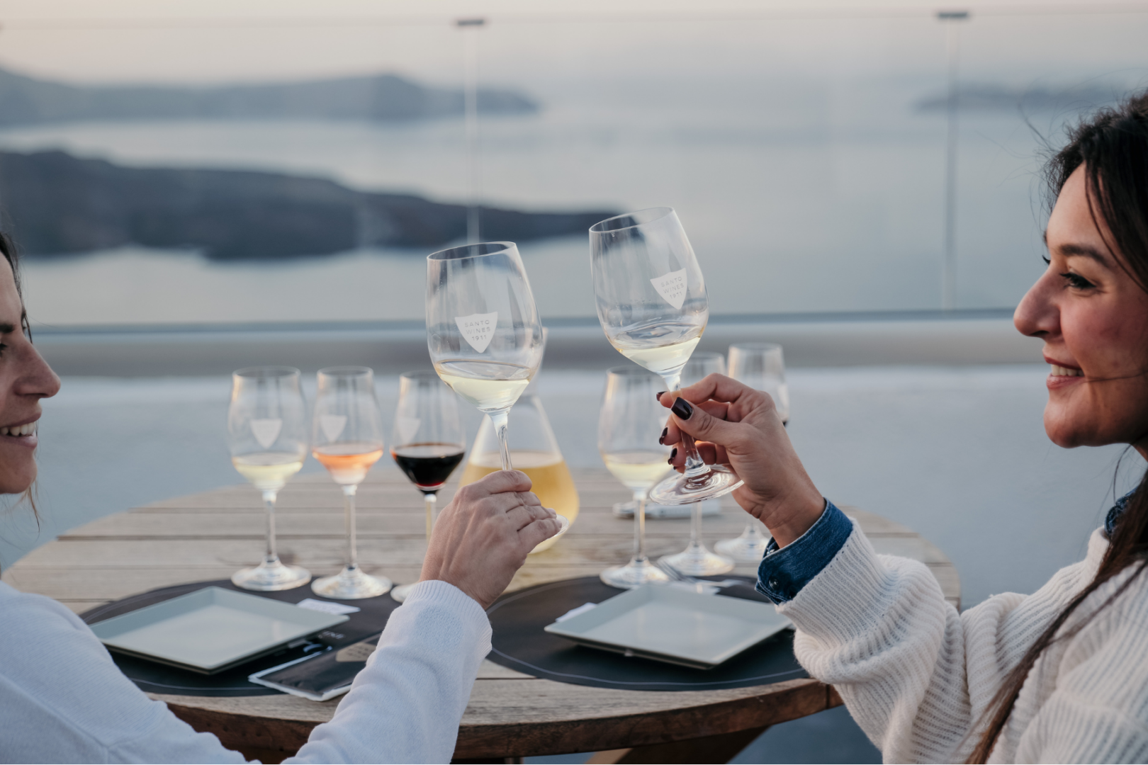 Wine Tasting with Caldera Views – Santo Wines Santorini Private Tour Two women toasting with Santo Wines glasses on a terrace overlooking the Santorini caldera, with a full wine tasting flight of white, rosé, red and sweet wines on the table.