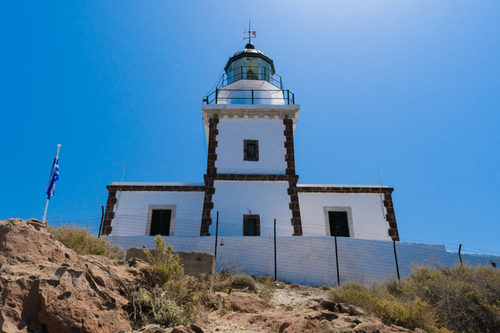 Akrotiri Lighthouse Santorini | Southernmost Point on the Private Island Tour Akrotiri Lighthouse Santorini with Greek flag against blue sky