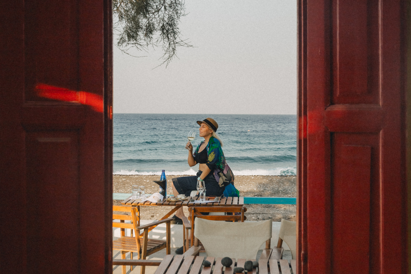 Beachside Wine Tasting in Santorini Woman sitting at a wooden table on a Santorini beach holding a glass of white wine, with the Aegean Sea visible through a red doorway frame