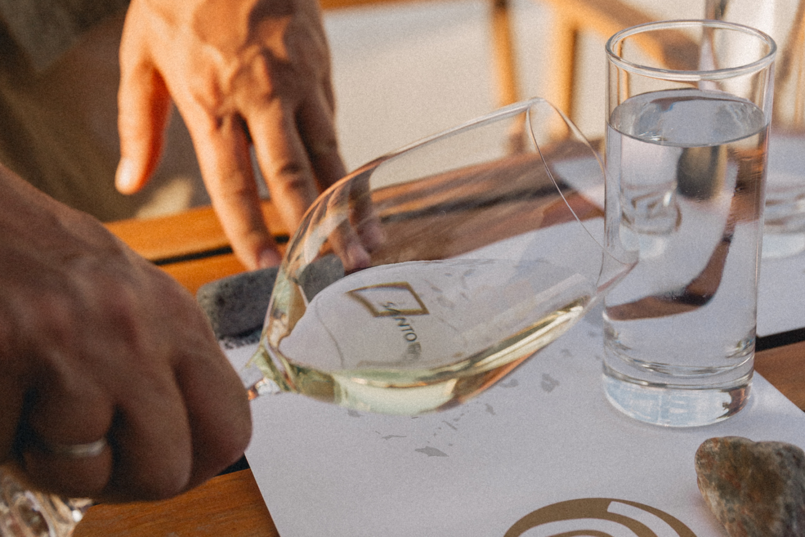 Santo Wines Tasting — Santorini Wine Tour Stop A Santo Wines branded glass being tilted and swirled on a tasting table with volcanic stones and a water glass beside it