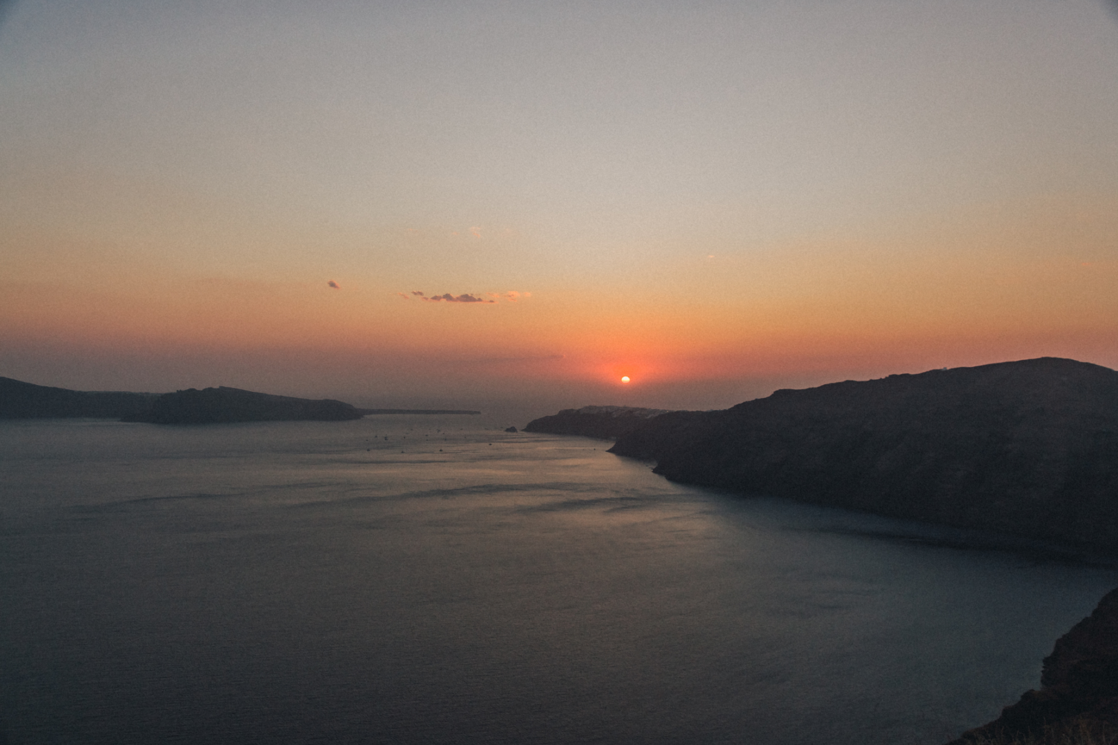 Santorini Sunset Over the Caldera — Dusk View Panoramic dusk view of the Santorini caldera with deep orange and pink sky reflected on the calm Aegean Sea between volcanic cliffs