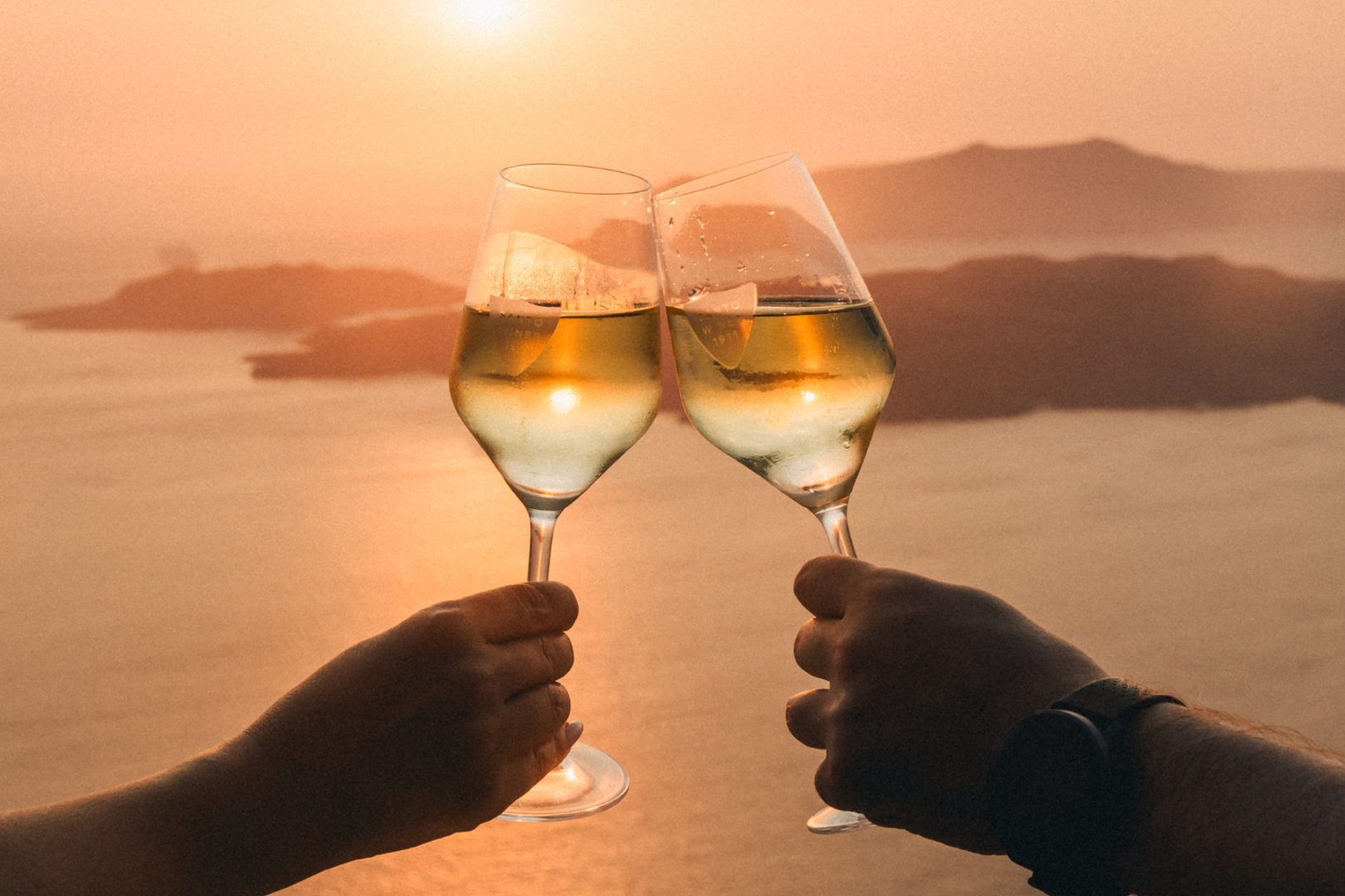 A Toast to Santorini — Private Sunset Wine Experience Two hands clinking glasses of white wine together in front of a glowing Santorini caldera sunset with volcanic islands in the background