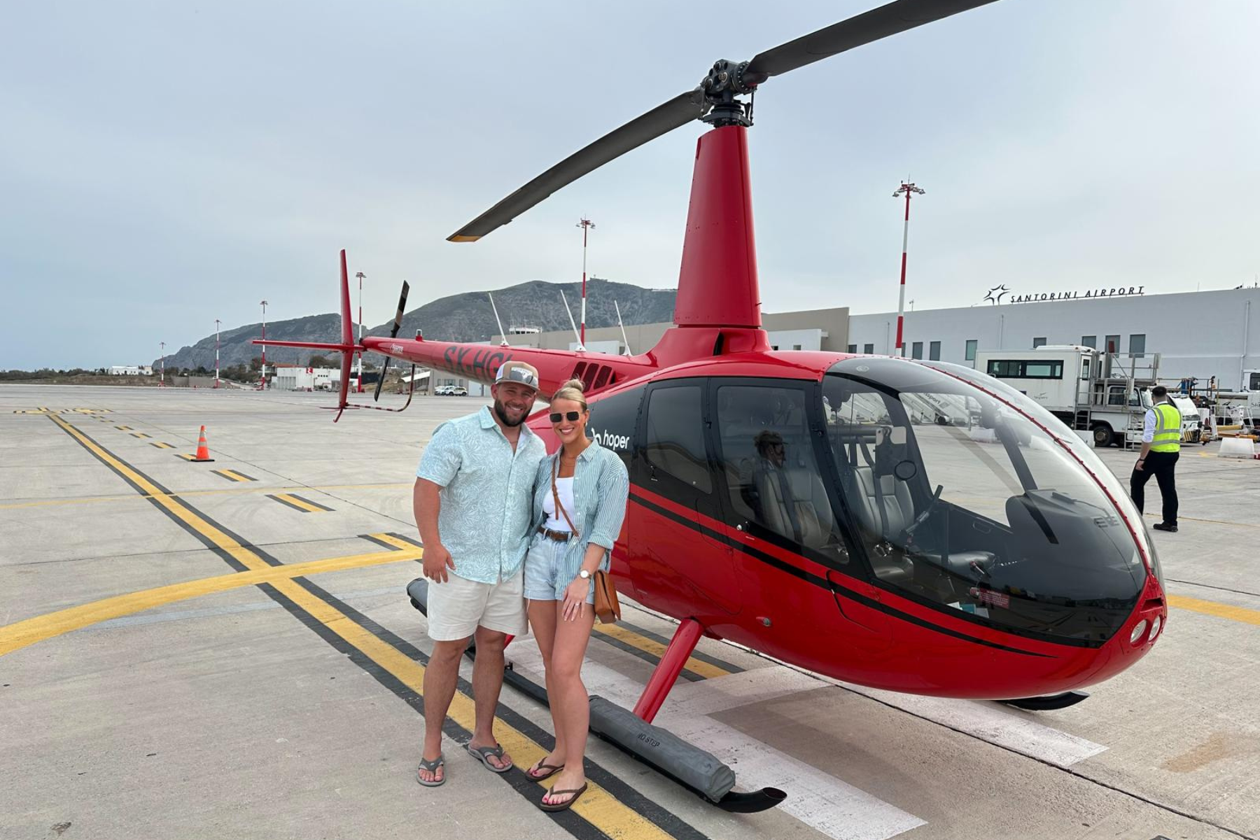 Santorini Helicopter Ride Two people stand proudly in front of a red helicopter at Santorini Airport, ready for a private sightseeing and wine tour.
