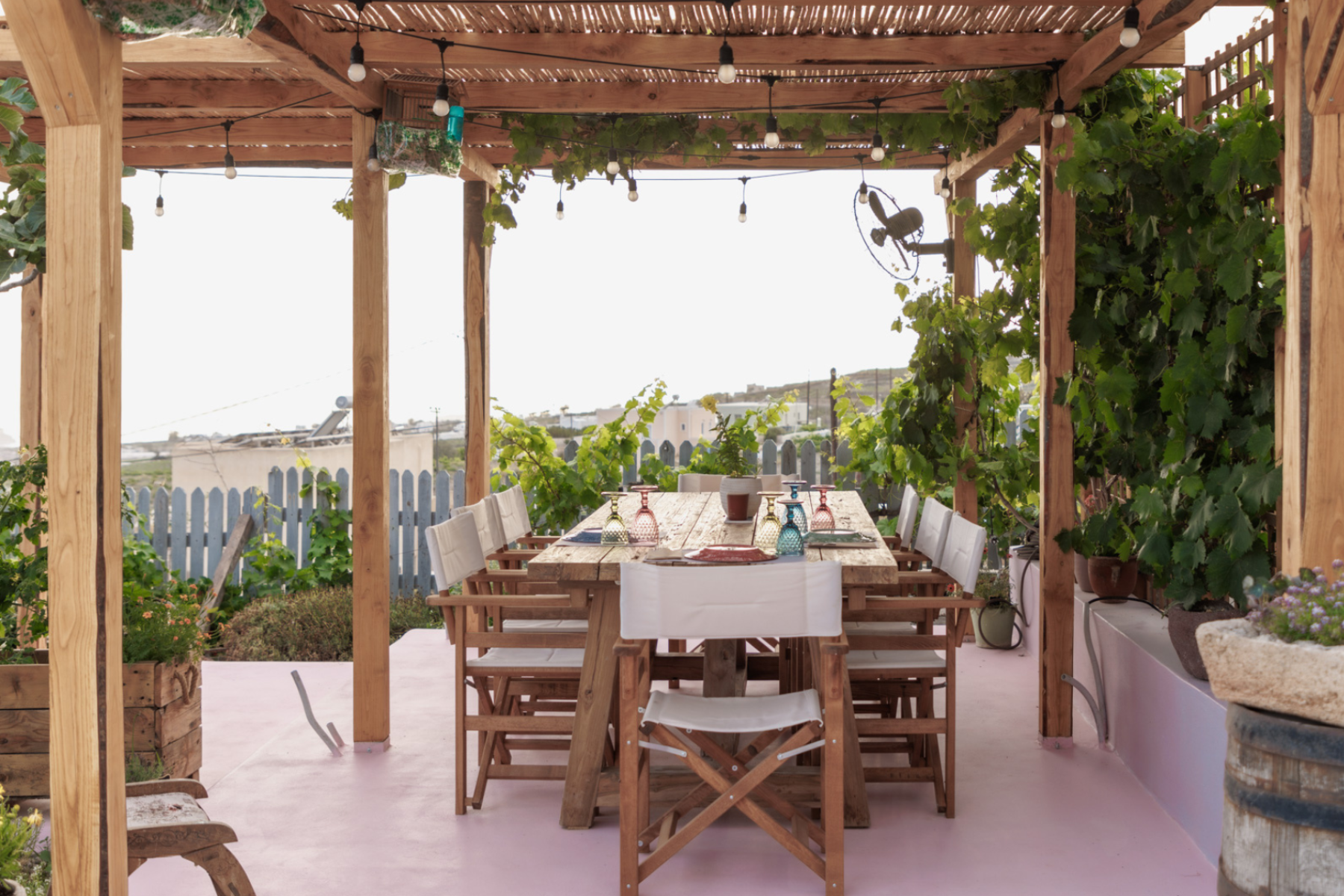Farm Outdoor Dining Space A sneak peek of the farm lunch, set under a wooden pergola surrounded by lush greenery, adorned with string lights and colorful glassware—perfect for concluding the Santorini Island Tour.