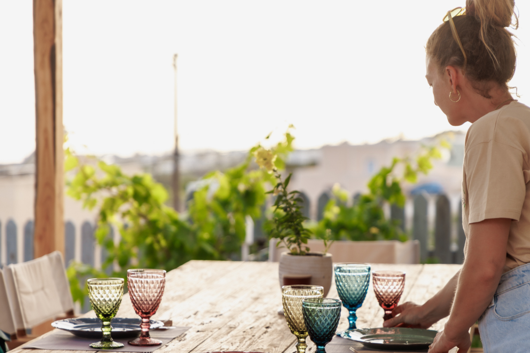 Setting the Scene for Outdoor Farm Dining A woman setting the stage for an outdoor farm dining table adorned with colorful glassware, framed by a backdrop of greenery and soft natural light