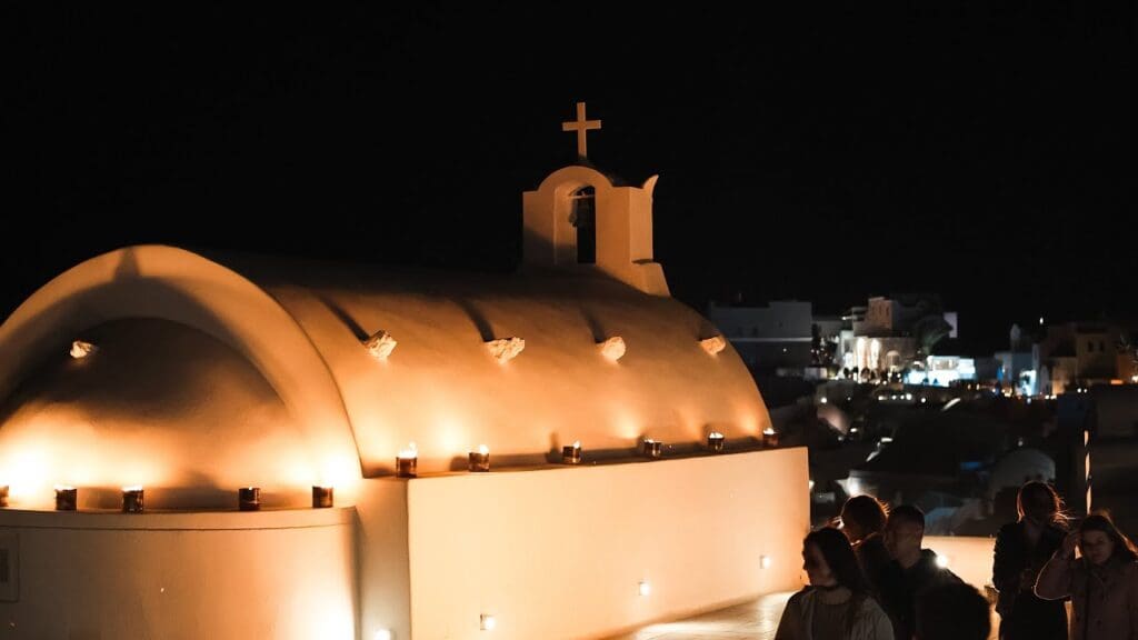 A whitewashed church in Pyrgos Kallistis, Santorini, illuminated by candlelight at night, with people observing in the background