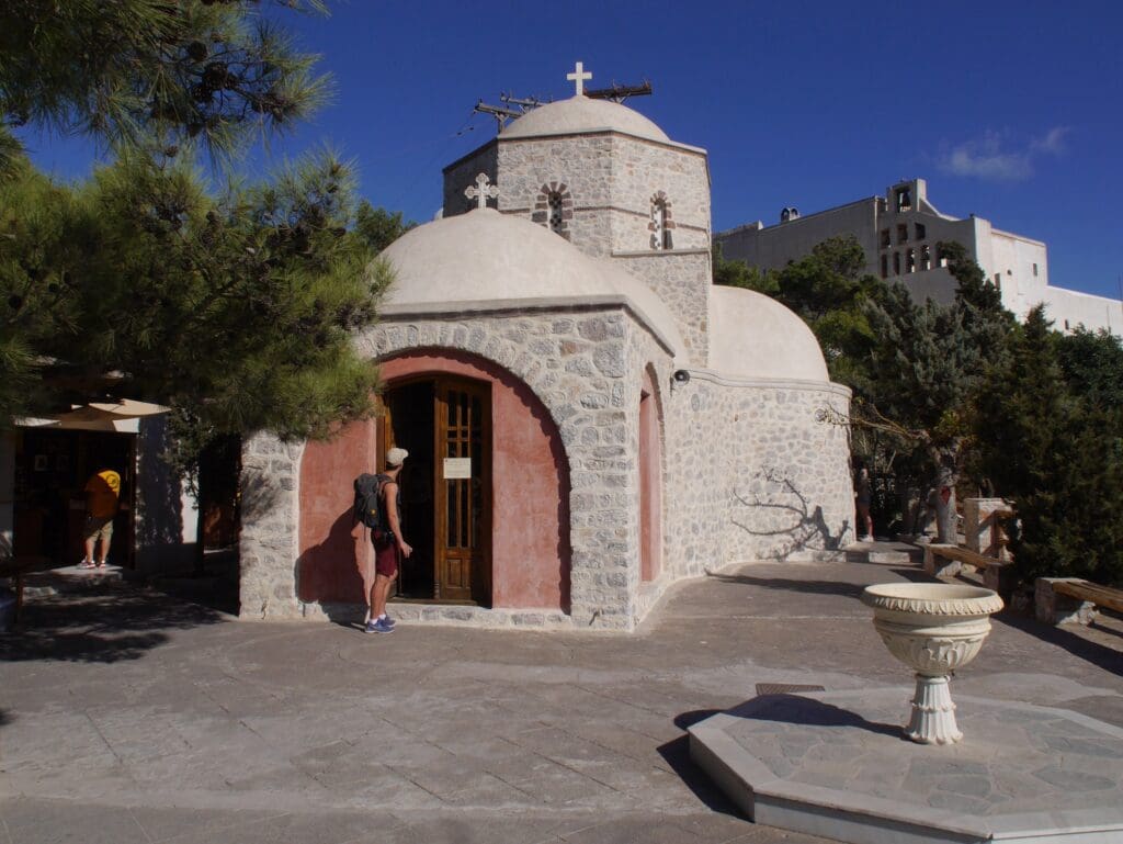 A stone church at Profitis Ilias Monastery in Santorini, Greece, with a person standing near the entrance. The scene is peaceful and serene.