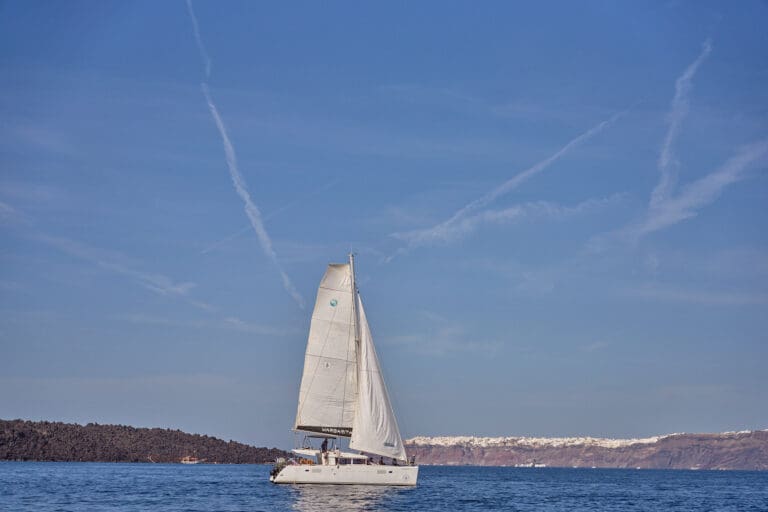 Sailing catamaran cruising near the coastline of Santorini with rugged cliffs and clear blue waters in the background.