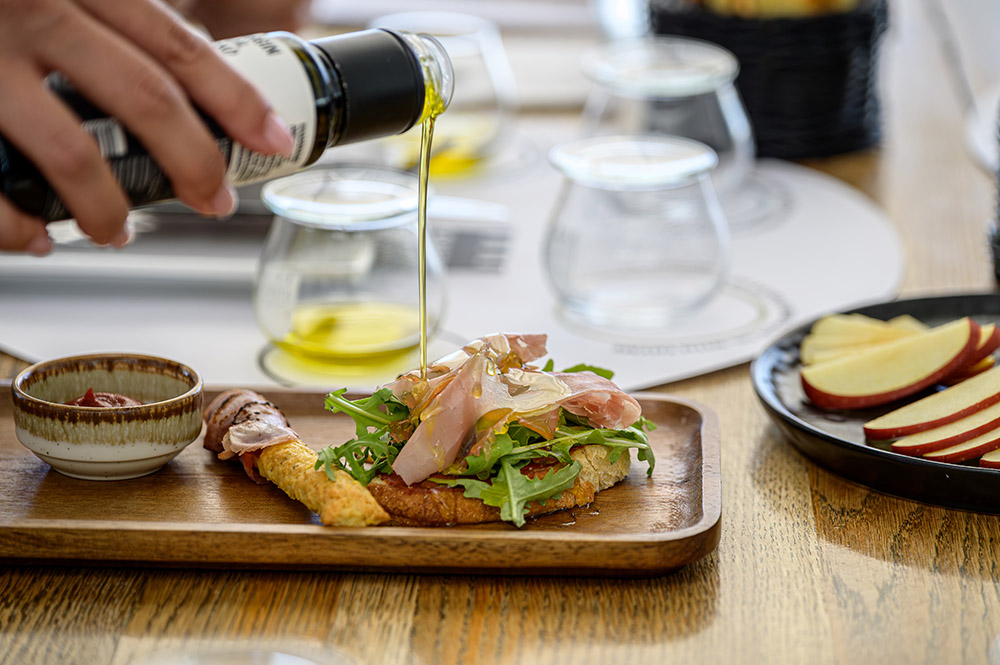 Santo Roots Olive Oil Drizzle Over Bruschetta – Olive Oil Tasting at Santo Wines Santorini Hand pouring Santo Roots extra virgin olive oil over a bruschetta with prosciutto and arugula on a wooden board, with olive oil tasting glasses in the background at Santo Wines, Santorini.