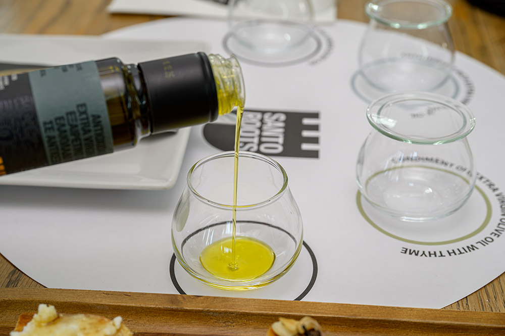 Santo Roots Extra Virgin Olive Oil Tasting Flight – Santo Wines Santorini Santo Roots extra virgin olive oil being poured into a glass tasting cup on a Santo Roots tasting mat, with empty tasting glasses and bread in the foreground at Santo Wines, Santorini.