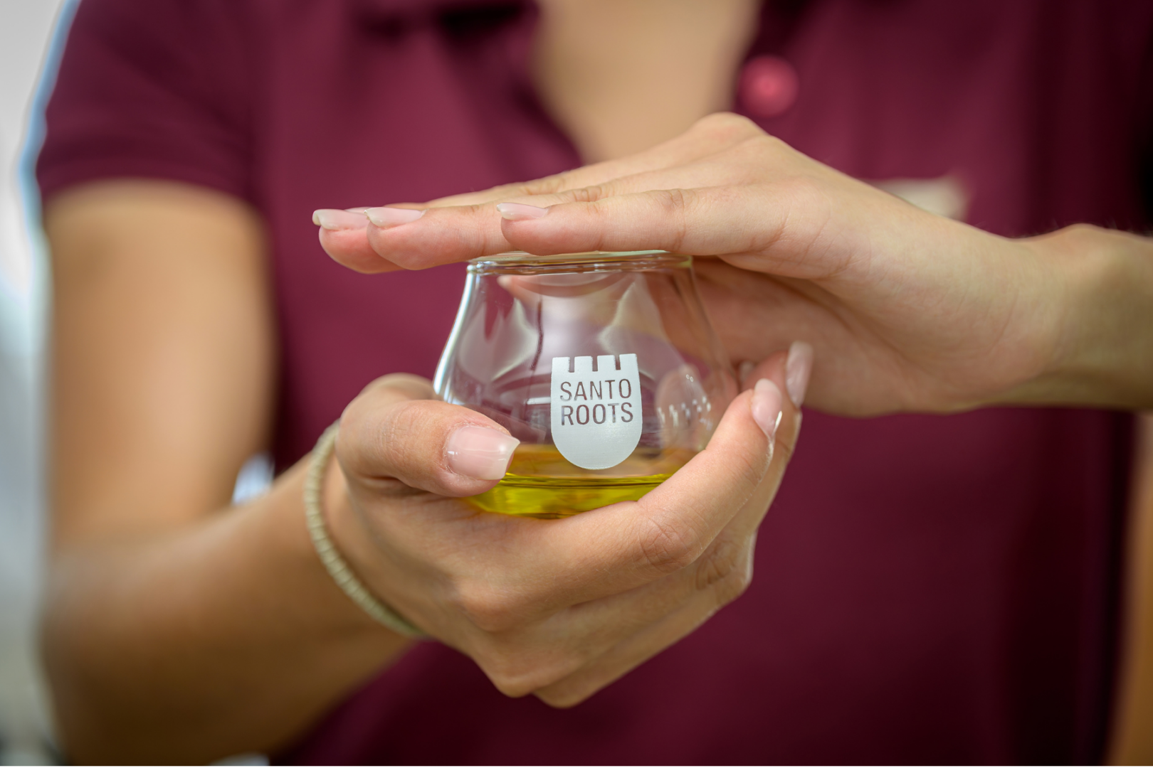 Santo Roots Olive Oil Tasting Glass – Expert Guided Experience Santorini Staff member in Santo Roots uniform warming a branded glass tasting cup filled with golden extra virgin olive oil in both hands.