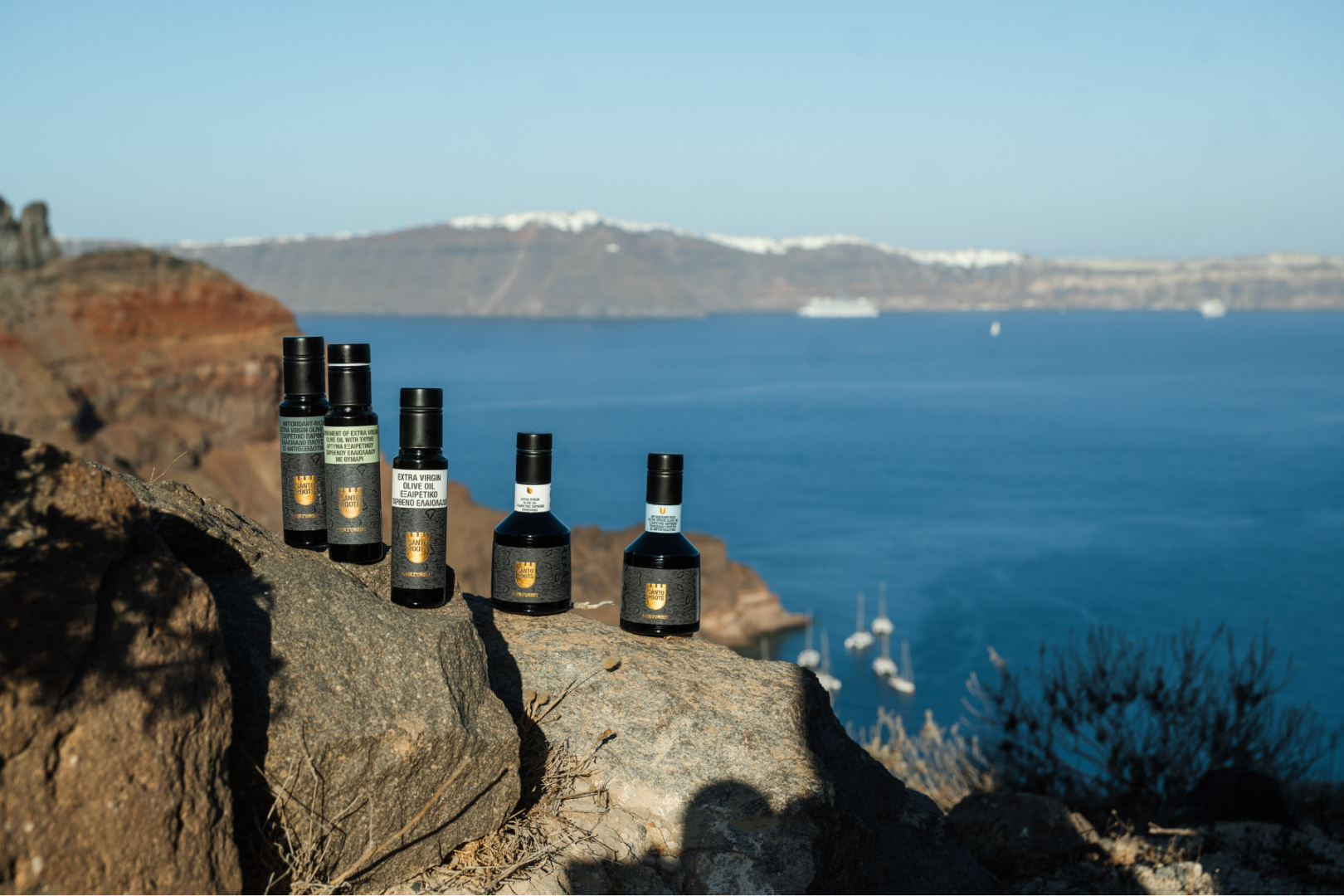 Santo Roots Olive Oil Collection – Santorini Caldera Views Five bottles of Santo Roots extra virgin olive oil and infused olive oil varieties lined up on volcanic rock with panoramic views of the Santorini caldera and Aegean Sea in the background.