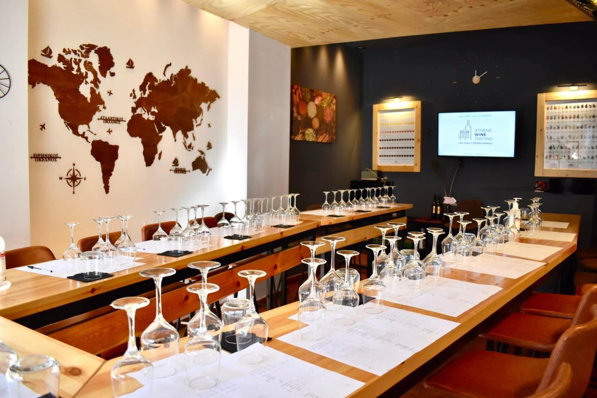 Athens Wine Tasting Experience Under Akropolis A room prepared for a wine tasting experience in Athens, Greece, featuring rows of empty wine glasses on long wooden tables. A large wooden world map hangs on the wall, and a screen displays "Athens Wine Tasting."