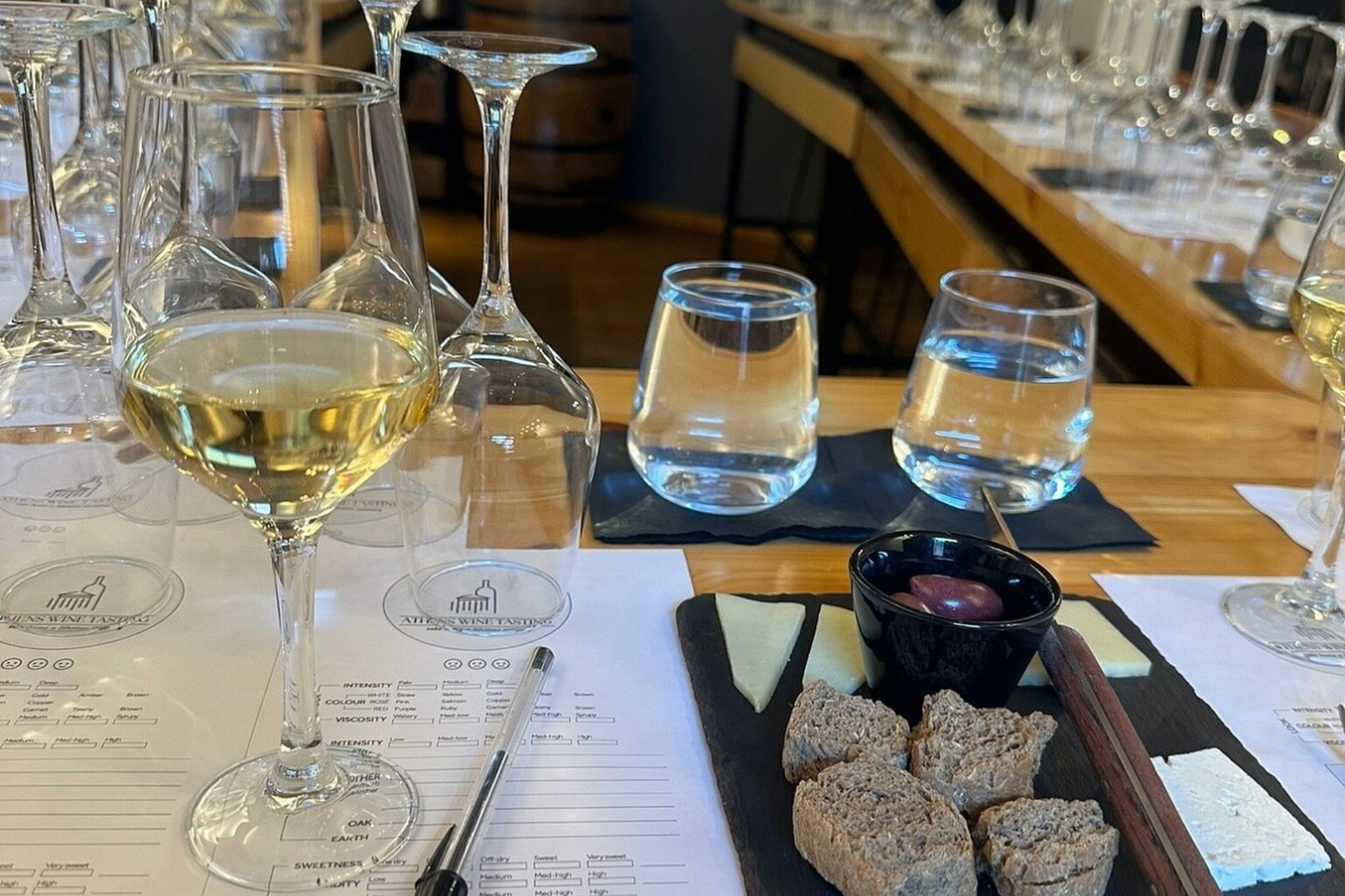 Athens Wine Tasting Experience Under Akropolis Wine glasses filled with wine and water are arranged on a table at the Athens Wine Tasting experience, accompanied by a slate board with cheese, bread, and olives. A pen lies next to a tasting sheet.