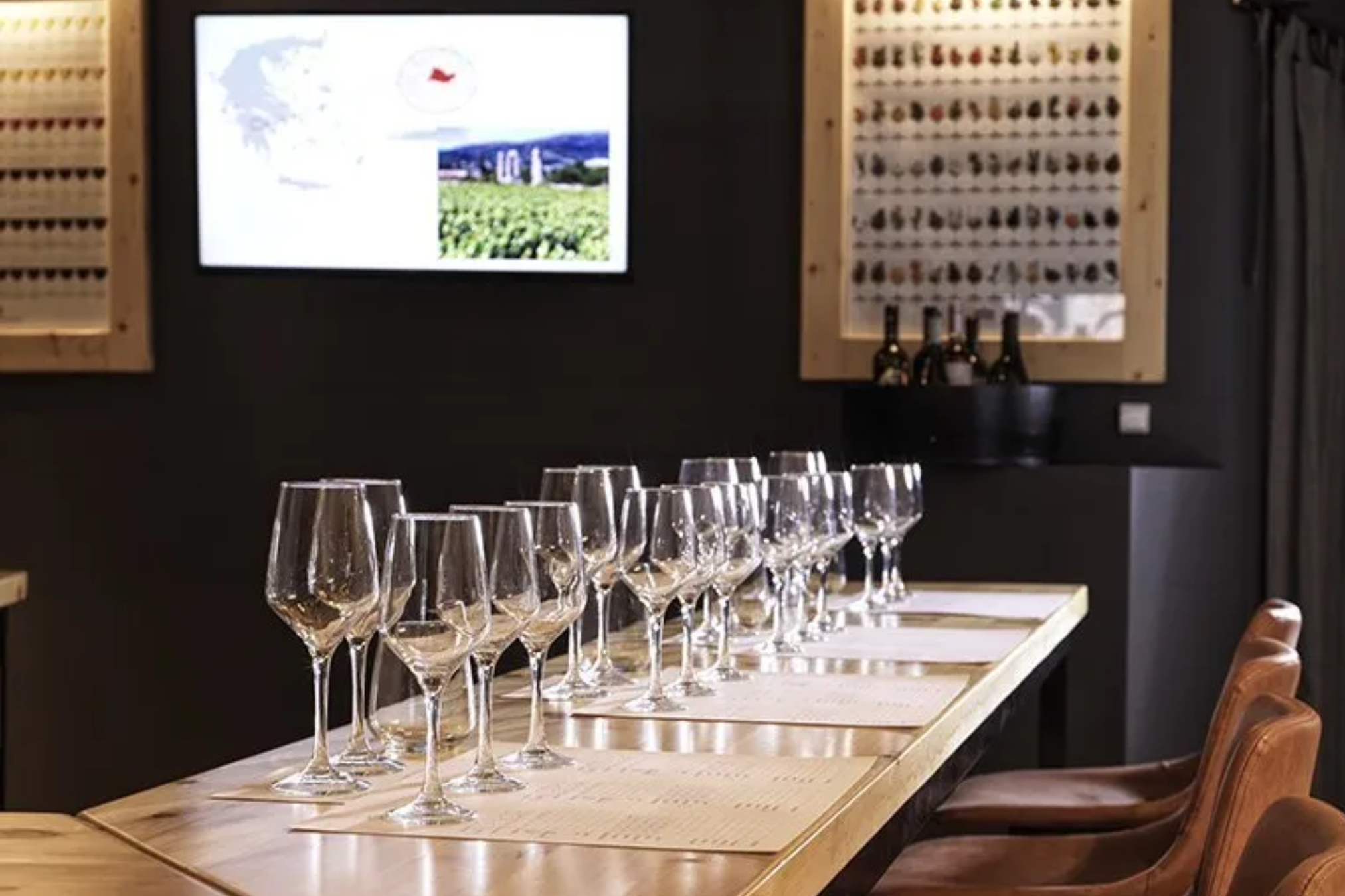 Ready for a Wine Tasting in Athens A long wooden table is set with wine glasses and tasting notes in preparation for a wine tasting experience. A screen displays a map of Greece and vineyard scenery.