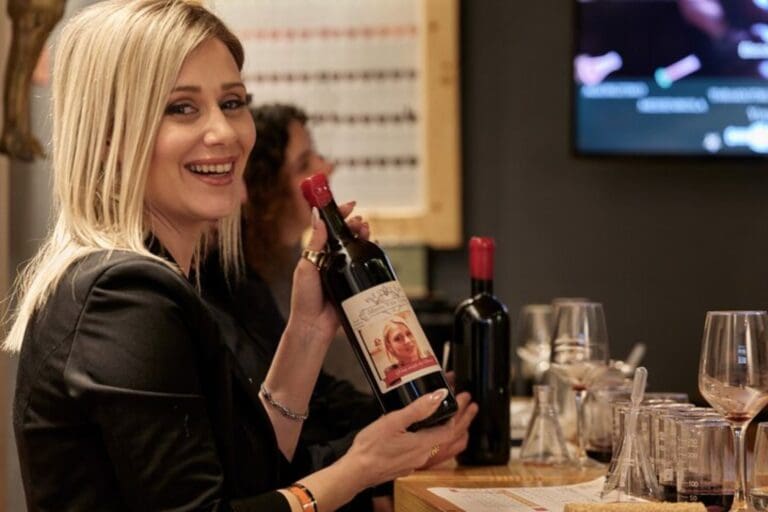 A smiling person holding a bottle of wine with a personalized label at a winemaking workshop. Wine glasses and another bottle of wine are on the table in front of them.