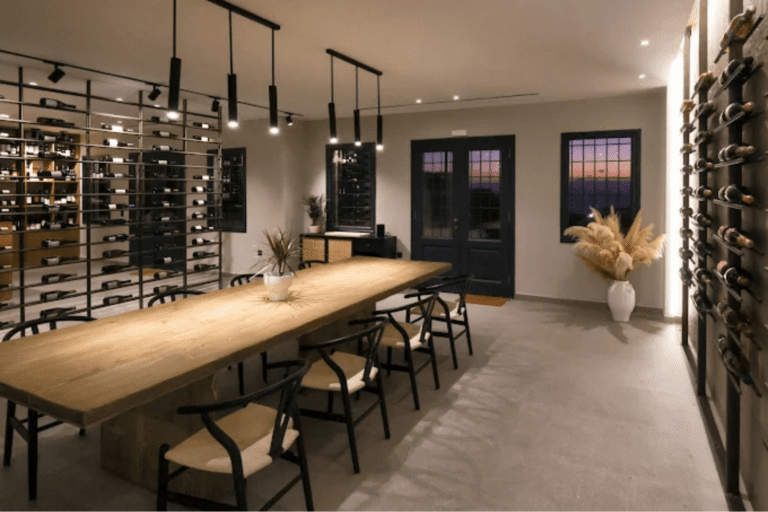 Interior of a local wine cava in Pyrgos Kallistis with a long wooden tasting table, black chairs, wall‑mounted wine bottle racks, and warm ambient lighting.
