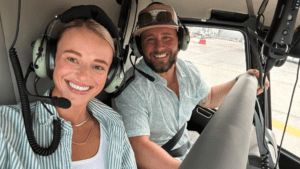 Two passengers wearing aviation headsets seated inside a helicopter cockpit on a runway, photographed from the front passenger area.