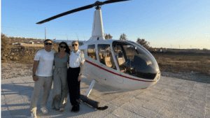 Three people standing beside a light helicopter parked on a paved helipad in an outdoor rural landscape.