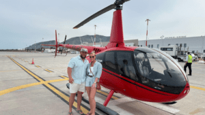 Two people standing beside a red light helicopter parked on an airport tarmac with terminal buildings visible in the background.