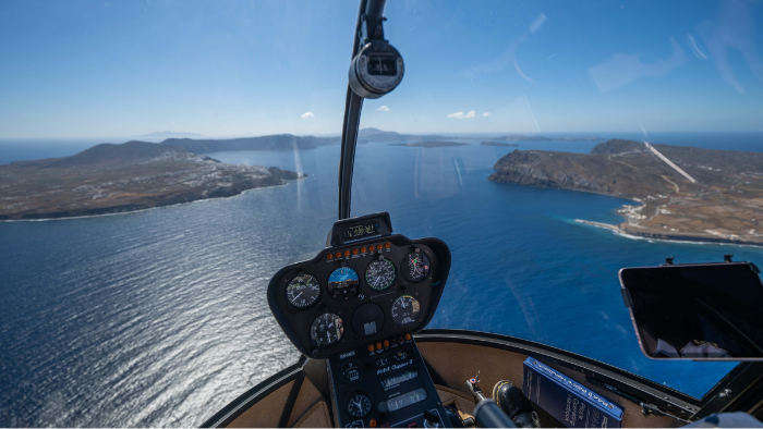 View from inside a helicopter cockpit overlooking the sea and coastal islands during flight, with flight instruments visible in the foreground.