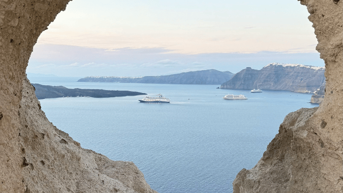Cruise ships anchored in the Santorini caldera viewed through a volcanic stone arch, a common sight during peak season