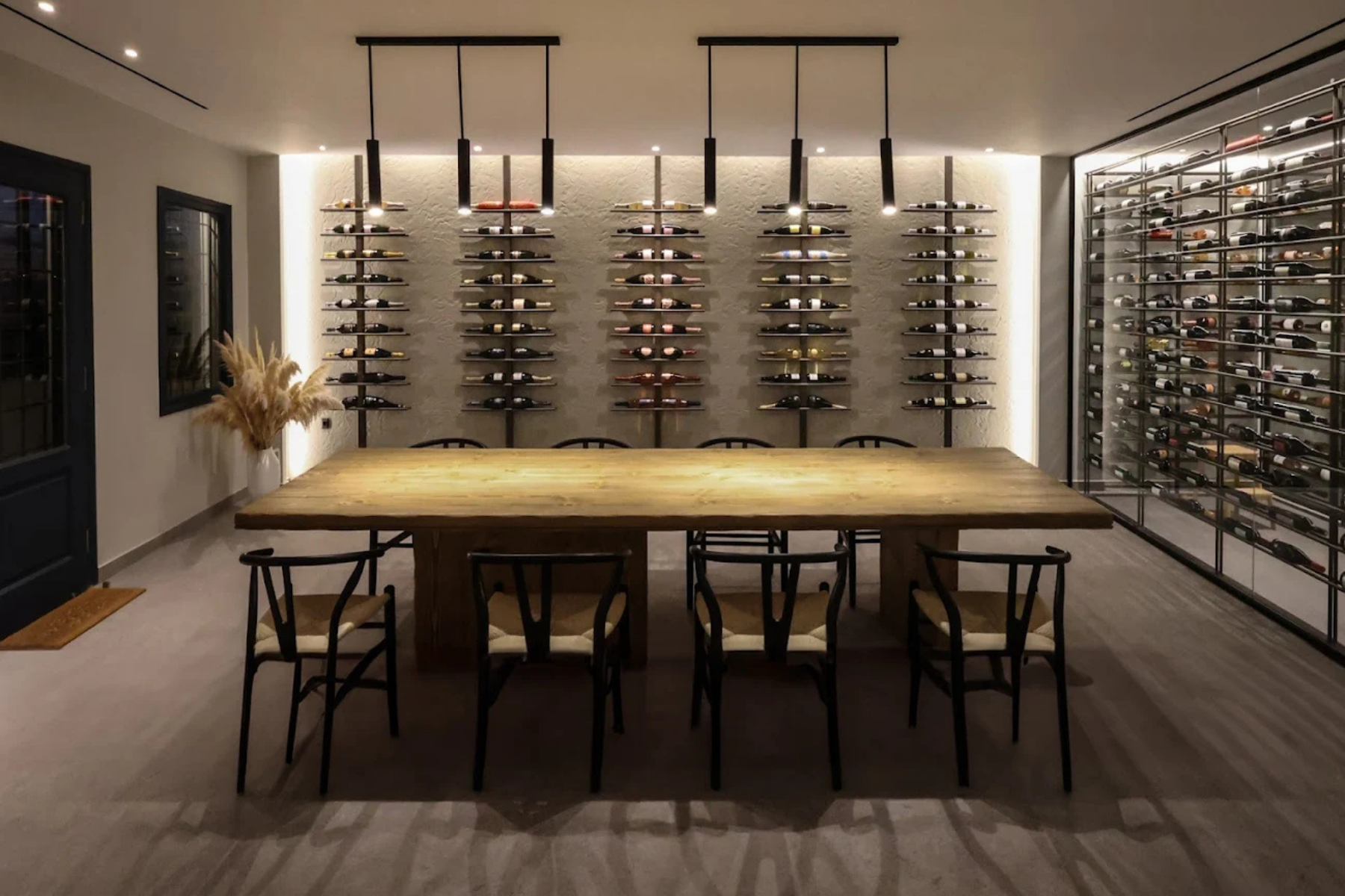 Local Wine Cava in Pyrgos Kallistis, Santorini Interior of the local wine cava in Pyrgos Kallistis with a long wooden tasting table, chairs, wall‑mounted wine bottle displays, and modern pendant lighting.