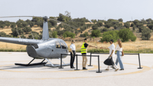 A light helicopter parked on a helipad with several people approaching and standing nearby in an open, rural landscape.