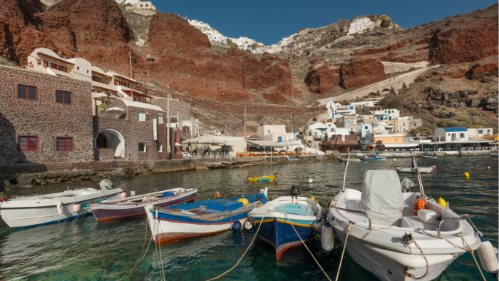 Colorful fishing boats moored in the calm harbor of Ammoudi Bay, Santorini, with traditional stone-built tavernas and red volcanic cliffs in the background