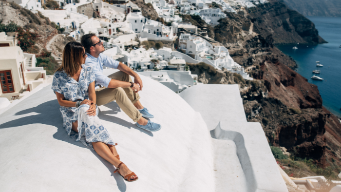 Two travelers sitting on a white Santorini rooftop overlooking Oia village and the caldera