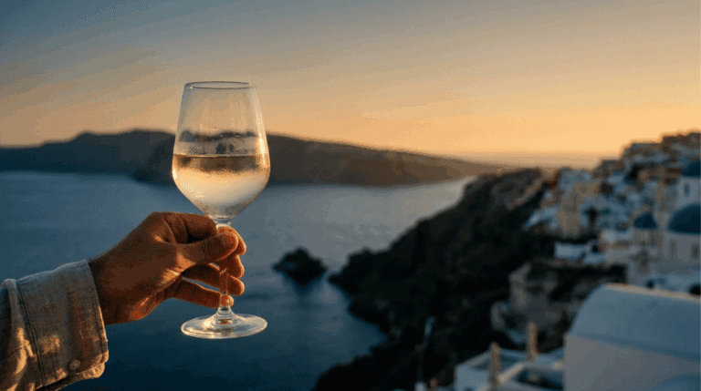 Wine glass of chilled Assyrtiko held against a Santorini caldera sunset, with white cliffside village in the background