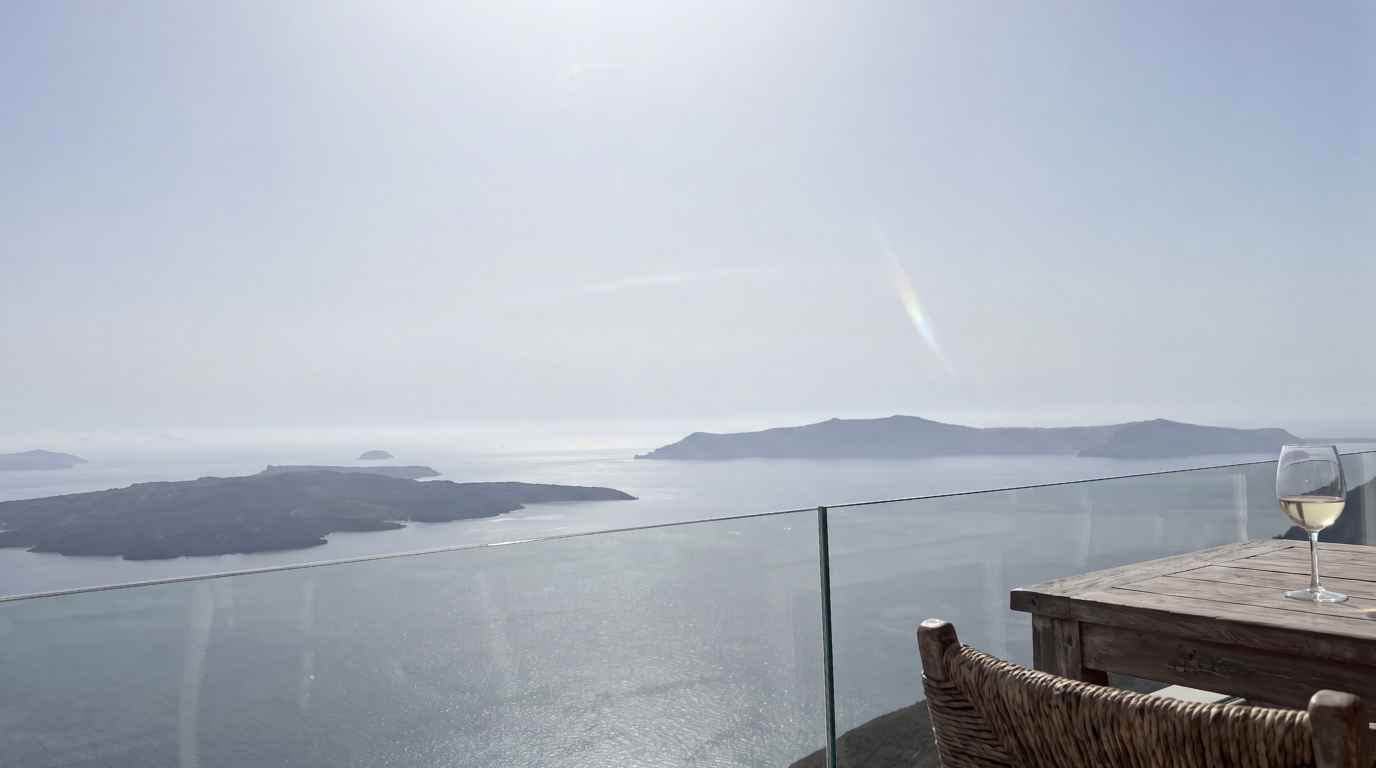 A glass of white wine on a terrace table overlooking the Santorini caldera and volcanic islands