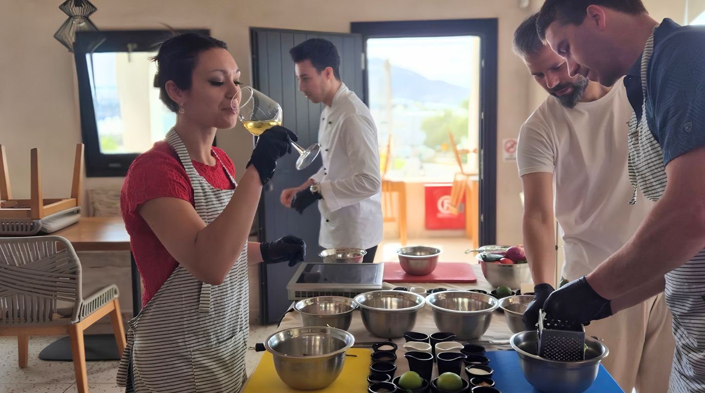 Guest tasting white wine while others prepare food, showing the pairing served alongside a Santorini cooking class